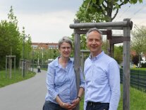 Eine Frau und eine Mann stehen vor einem Baum an einer frisch gepflanzten Baumallee. 