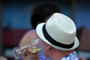 Kopf eines Mannes von seitlich hinten mit hellem Hut, der ein Glas mit Weißwein zum Mund führt.