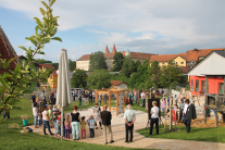 Blick auf eine Stadtsilhouette mit Kirchtürmen und alten Bäumen. Davor ein neu gestalteter Spielplatz, auf dem Kinder und Erwachsene versammelt stehen. 