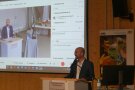 Jörg Hirsche steht am Rednerpult mit Livesendung per Video im Hintergrund