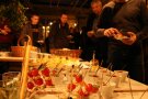 Auf einem Tisch sind verschiedene Häppchen angerichtet, unter anderem Mozzarellakugeln und Cocktailtomaten auf Schaschlikspießen. Bei gedämpftem, gelbichem Licht bedienen sich einige Personen an der Speisenauswahl.