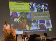 Wolfgang Palme steht am Rednerpult, im Hintergrund in der Präsentation seine beiden Bücher zu Wintergemüse mit Bildern zu Gemüse im Winter, darauf Lauch, Karotten, Rüben und auch Personen.