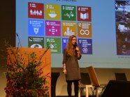 Katrina Schwanke steht neben dem Pult, im Hintergrund auf der Leinwand sind zwölf Symbole der UN-Charta der Nachhaltigkeit.