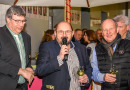 Drei Herren halten ein Weinglas in der Hand, während der mittlere in ein Mikrofon spricht.