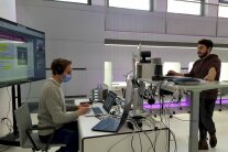 Referent am Stehtisch, Techniker sitzt am Tisch, viele Kabel, viel Technik, großer Bildschirm