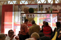 mehrere Menschen stehen an einer Weinbar, im Vordergrund wird gegessen und getrunken